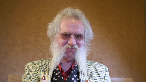 Older man with beard strokes his hair while shaking his head at the camera