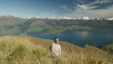 Blond-woman-relaxing-in-grass-looking-at-Southern-Alps-with-snowy-peaks-and-Lake-Wanaka