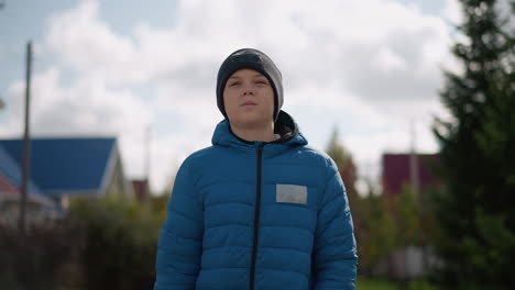 close-up of white boy in blue jacket and black beanie strolling in residential area with a frowning face, reflecting subtle emotions against a backdrop of autumn colors