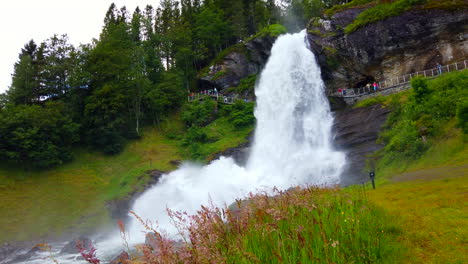 Dramatic-Steinsdalsfossen-Waterfall-Steine,-Norway-wide-from-front---pan-left-through-flowers--you-can-see-the-walkway-and-people-behind-the-falls,-4k-ProRezHQ