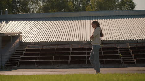 young girl in hoodie and jeans with long hair tied back walks pensively near empty stadium seating on a sunny day, arms folded, she appears reflective, surrounded by empty bleachers and greenery