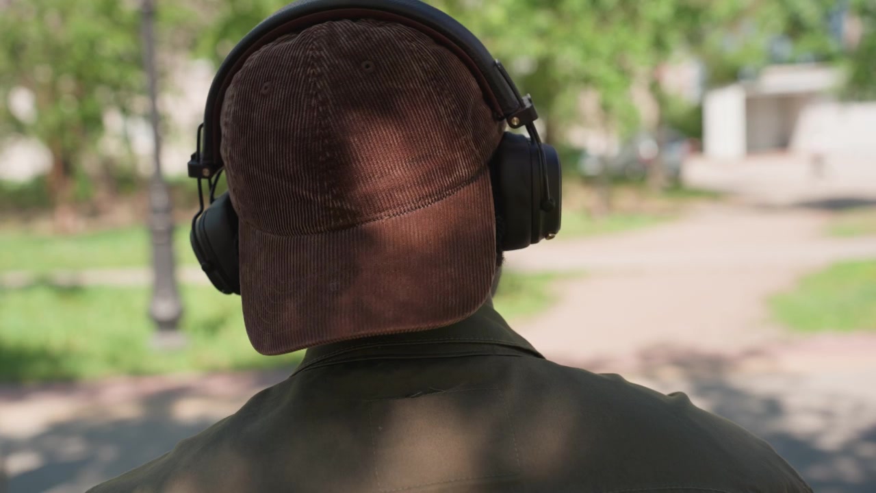 Primo piano di un uomo con berretto marrone e cuffie in un parco con pedoni sfocati che camminano. L'uomo rimane fermo mentre le persone si muovono sullo sfondo creando un senso di ambiente pubblico