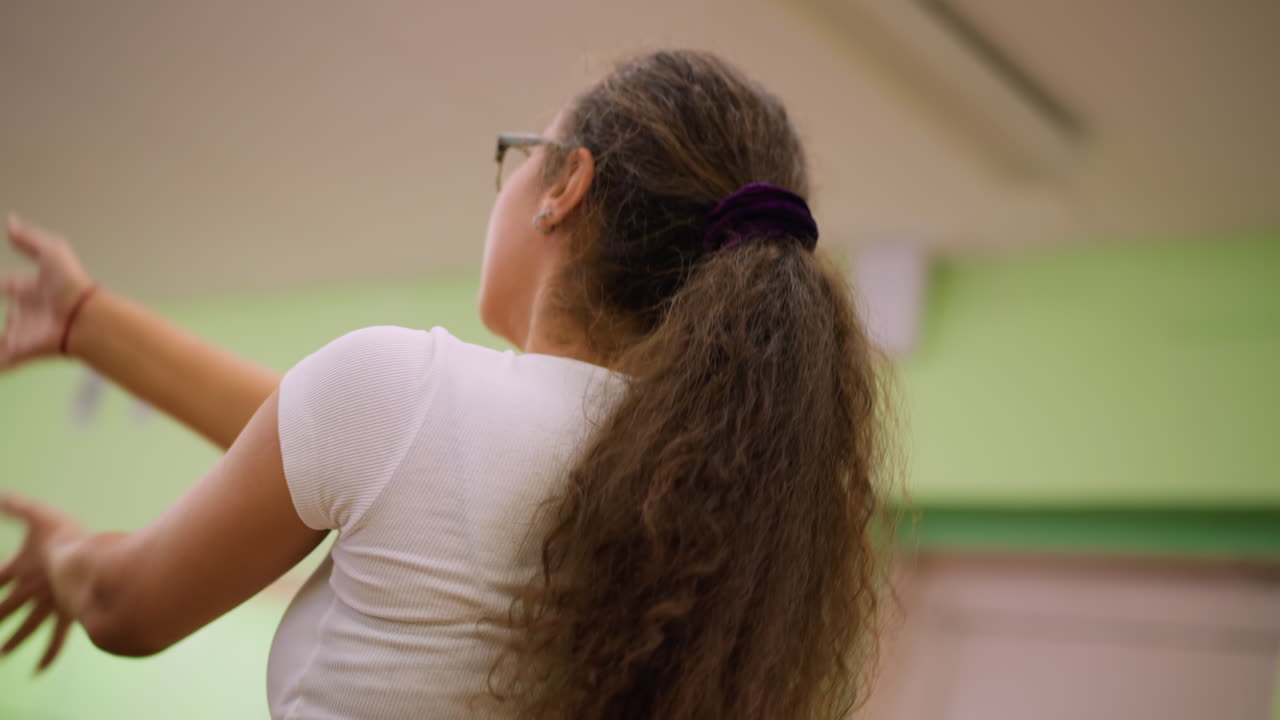 Side view of woman in white top and eyeglass pushing chest outward, stretching hands and flipping hair with expressive movement during energetic indoor workout, highlighting strength, grace
