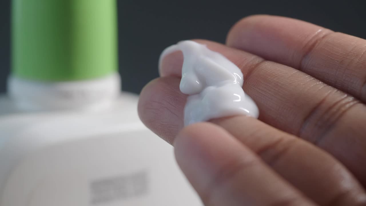 A hand with a dollop of white cream on the finger, with a bottle in the background