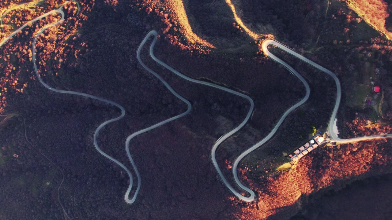 Winding mountain road forms a long series of linked hairpins across a dark leafless slope while patches of afternoon sun mark the ridges and a small roadside cluster remains at the far edge
