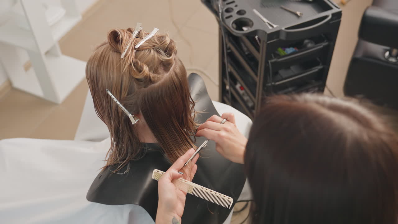Primer plano de una mujer negra sentada mientras una estilista le trenza el pelo y una ayudante observa, con detalle de las manos separando el pelo con clips y un peine, un carro de peluquería con productos, un interior moderno y luminoso, ambiente tranquilo y concentrado