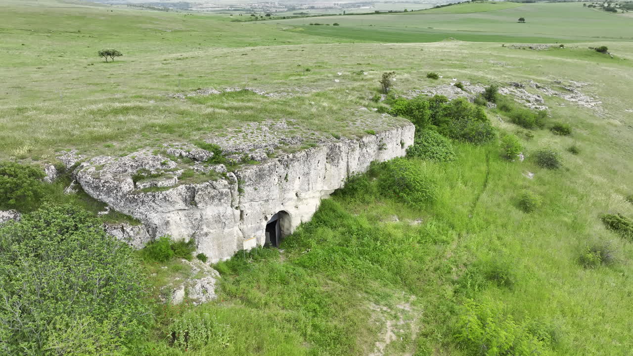 Оrbit drone view of a rock-hewn cave church set in lush spring greenery; limestone cliff façade with arched entrance, surrounding meadows and rolling fields under soft clouds
