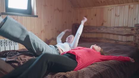 two joyful young women fall backward onto bed in cozy wooden room while laughing and holding hands, warm sunlight through window