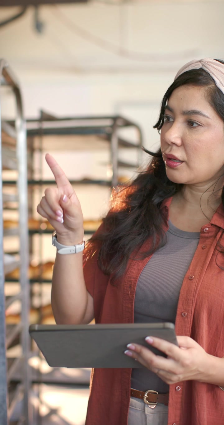 Video verticale: Donna di mezza età che legge un tablet, controlla gli scaffali e aggiorna l'inventario in una panetteria.