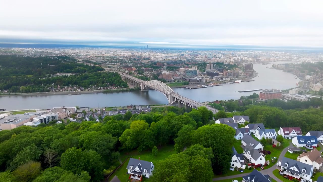 Vista elevada expansiva mostra um rio calmo serpenteando por uma cidade densa, ponte em arco de aço ligando ambas as margens, encosta verde vibrante em primeiro plano e casas brancas agrupadas à beira da água sob uma luz suave
