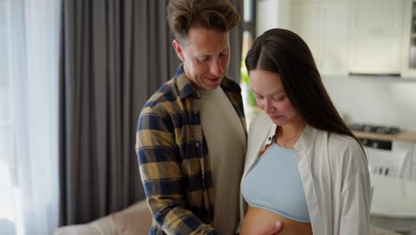 Happy-man-in-a-plaid-shirt-stands-near-his-pregnant-Brunette-wife-and-strokes-her-belly-while-relaxing-and-spending-time-in-a-modern-apartment