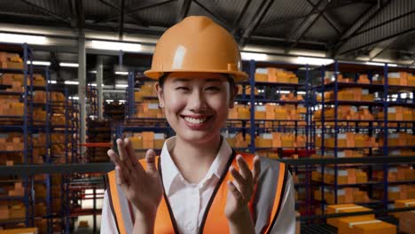 cerca de la ingeniera asiática con casco de seguridad de pie en el almacén con estantes llenos de productos de entrega. sonriendo a la cámara y aplaudiendo en el almacenamiento
