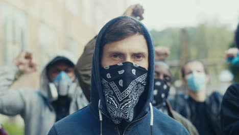 portrait of young handsome caucasian man in hood and scarf on mask looking at camera while standing in crowd of pretesters. close up of masked male face oudoor at strike.