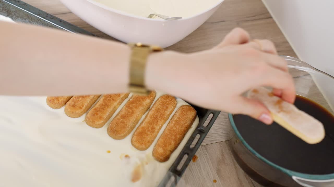 Close-up of hand placing golden ladyfingers on creamy mascarpone base, with bowl of coffee for soaking and utensils nearby, during tiramisu preparation on wooden kitchen surface