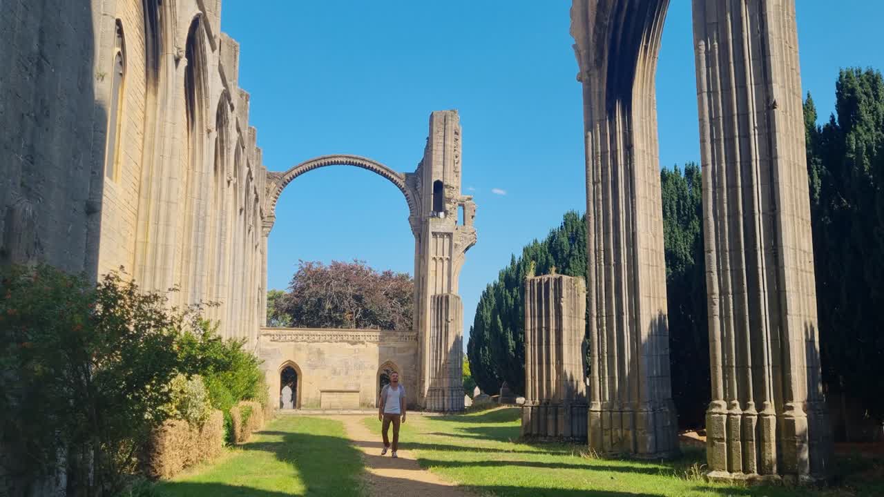 A male visitor wanders through the open-air ruins of Crowland Abbey, admiring centuries-old stonework beneath a bright blue summer sky in England