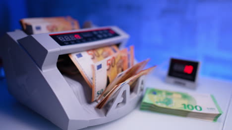 hand places a pile of orange euro banknotes into cash counting machine and starts working. woman takes the counted stack and puts money on the desk. close up. blurred backdrop.