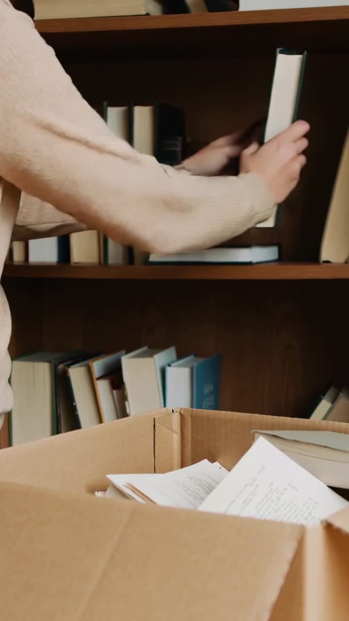 Video verticale: Donna con maglione beige che raggiunge una scatola, sollevando uno scaffale porta-libri verde scuro mentre disimballa a casa.