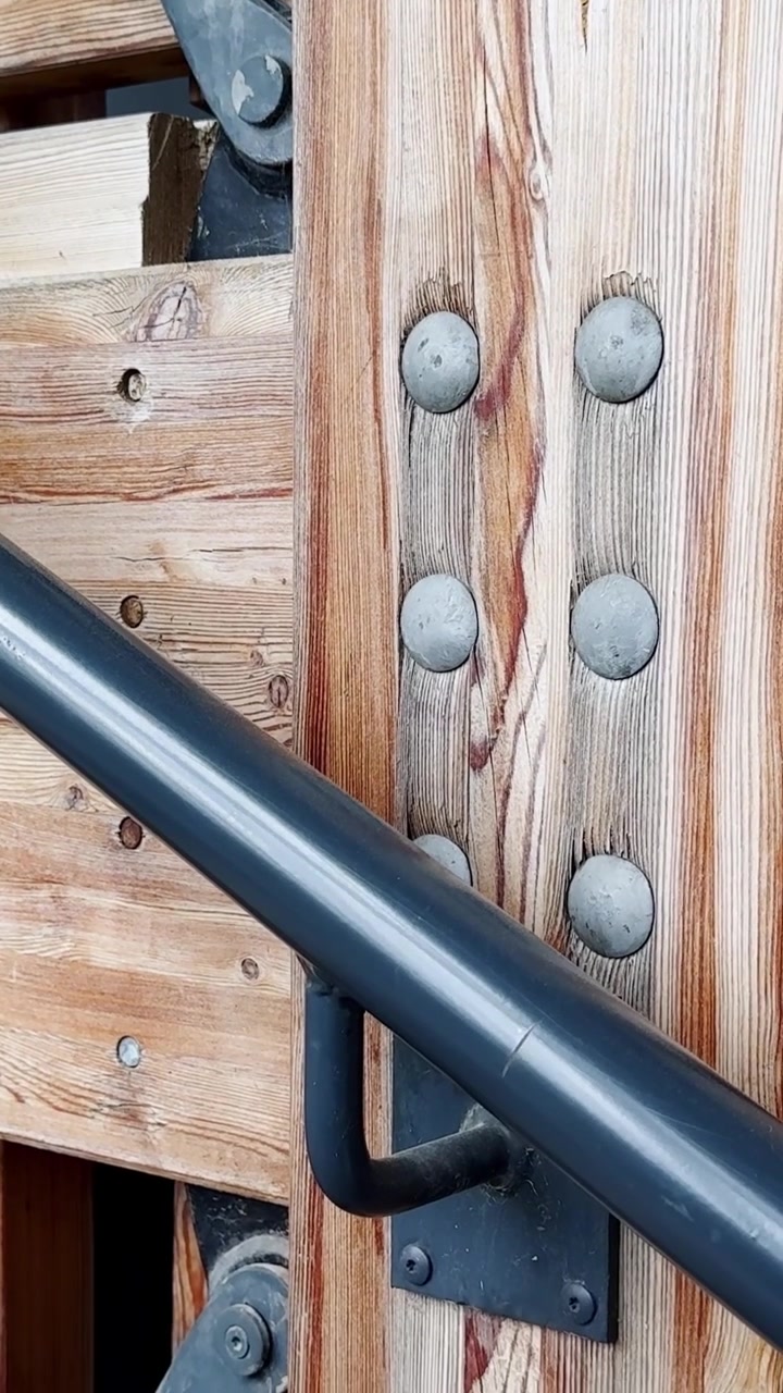 Primo piano di una ringhiera di scale in legno con corrimano in metallo, bulloni e giunti strutturali, che mostra dettagli di costruzione esterna e la venatura naturale del legno.