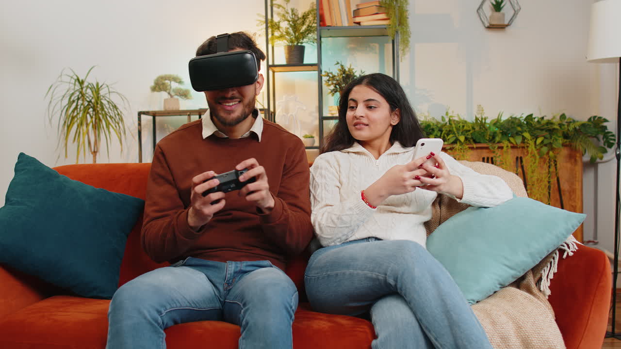 joven feliz con gafas vr jugando a videojuegos con joystick sentado al lado de su novia india