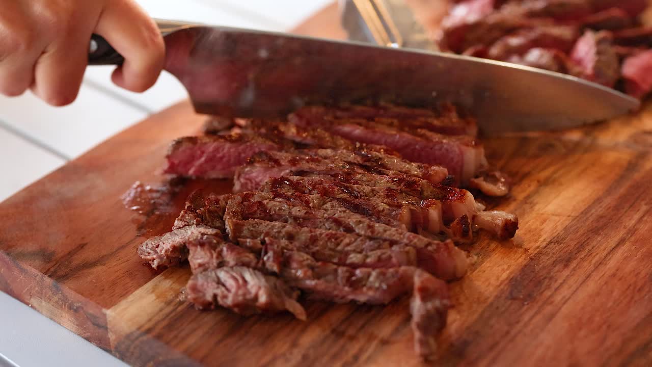 A chef expertly slices cooked beef steak on a wooden board under natural lighting in a kitchen environment