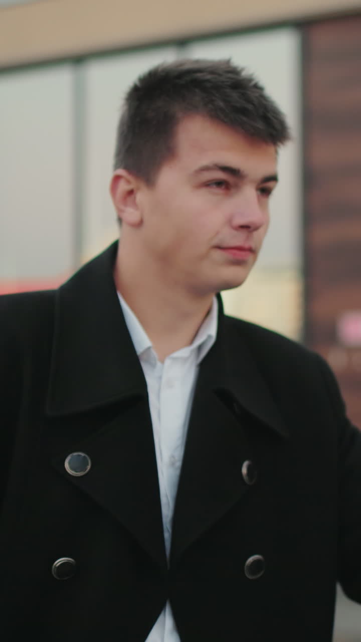 검은 코트를 입은 남자가 자신감있는 표정으로 시간을 확인하고, 주머니에 손을 넣고, 현대적인 유리 건물 앞에 서 있으며, 성공한 사업가는 도시 환경에서 전문성을 발휘합니다.