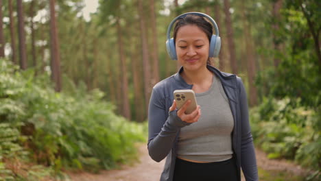 woman wearing wireless headphones exercising doing work out outdoors streaming music or podcast from mobile phone running along track in forest wearing sports clothing shot in real time