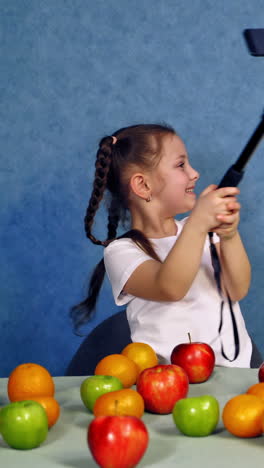 little girl doing selfie. cute child smiling to camera of the phone and showing tongue. fresh fruit on the table and a funny girl photographing. vertical video
