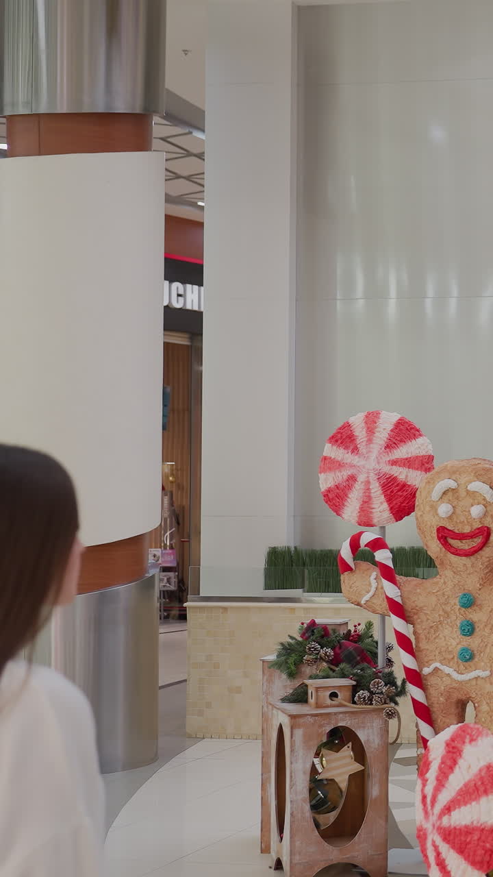 Back view of woman walking towards festive holiday tree with, Christmas decorations inside modern shopping mall, beautiful ornaments, candy canes, and gingerbread man near Christmas tree