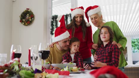 familia caucásica feliz de varias generaciones con sombreros de santa mirando el teléfono inteligente