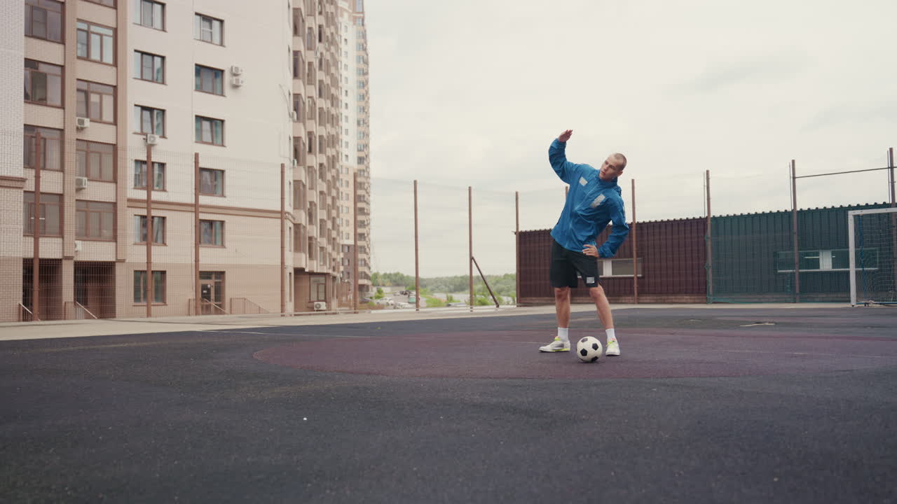 young man juggling football on court, rain drizzling across asphalt surrounded by apartment blocks and chainlink fence, dynamic footwork and freestyle tricks, intense concentration and gritty urban