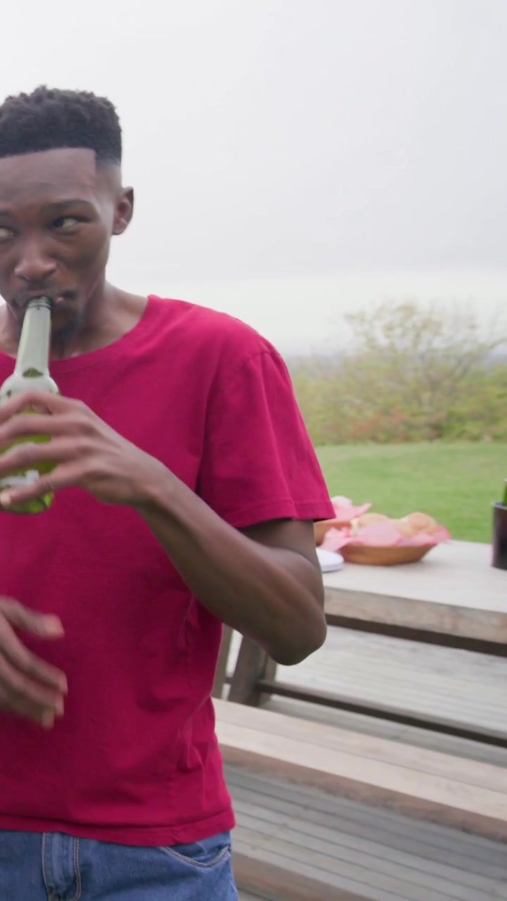 Vertical video: Bringing green bottles red tee man drinking on deck by cooler for meal, copy space