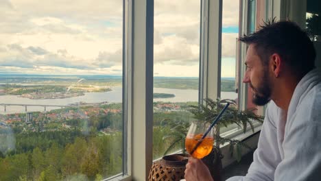 close-up of a man in a bathrobe sipping a drink, enjoying the view over sundsvall’s cityscape and port after a relaxing spa day. a quiet moment of wellness and reflection
