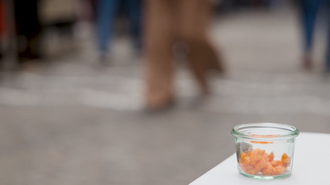 Glass jar of orange snack on table, pedestrians walking past, shallow focus, natural daylight