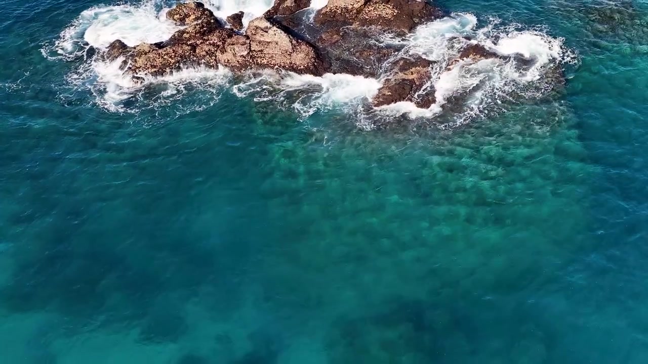 Ripresa aerea dall'alto di acqua oceanica turchese limpida e onde che si infrangono contro le rocce costiere