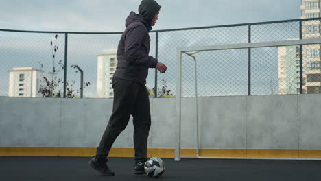 man practicing soccer drills on sport arena, skillfully pushing ball while appearing fatigued, goalpost and urban residential buildings in background