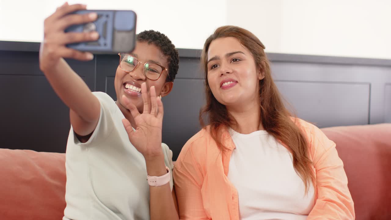 Amiche afroamericane sul divano che tengono lo smartphone angolandolo e salutando in chiamata