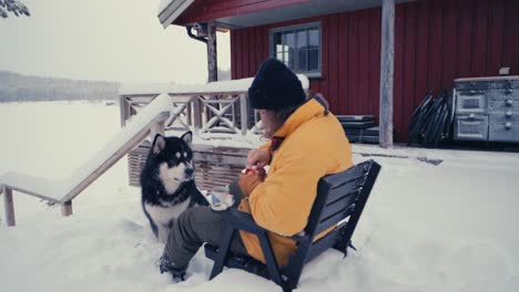 man sitting on chair and feeding his alaskan malamute dog while sitting on chair outside his cabin