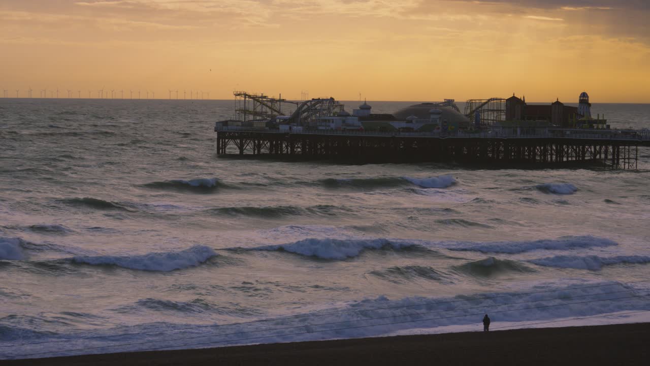 Brighton Palace Pier with Sunset Rays in Deep Background with Strong Sea Waves Crashing on Shoreline. Historic Building Landmark with Rides and Amusements in Sussex, UK