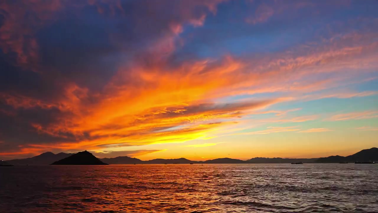 暗い海の上に小さなシルエットの島があり、鮮やかなオレンジ、赤、青の燃えるような雲が広がる息をのむような夕暮れのビデオ