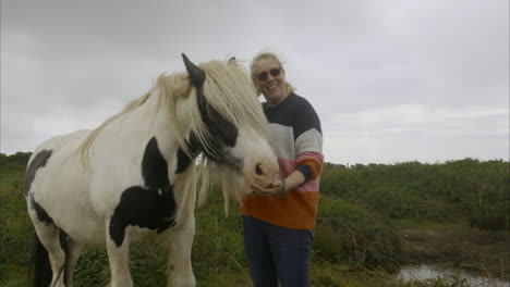 Beautiful-woman-beside-Piebald-horse-petting---posing-letting-long-hair-down,-wide-shot