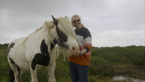 beautiful woman beside piebald horse petting - posing letting long hair down, wide shot