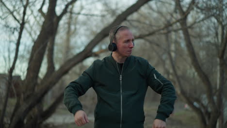 a man in a green jacket and headphones dances with expressive movements in a wooded area