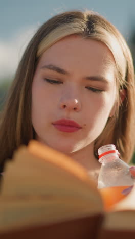mujer sentada al aire libre, sosteniendo un libro en una mano mientras toma un sorbo de una botella de jugo con la otra, el libro está ligeramente borroso mientras continúa leyendo, el fondo está borroso