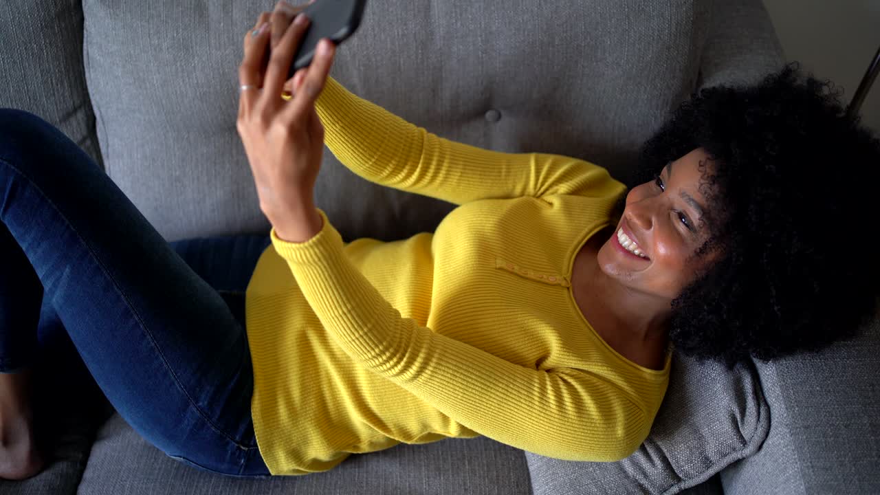 Positive black woman taking selfie with smartphone on sofa
