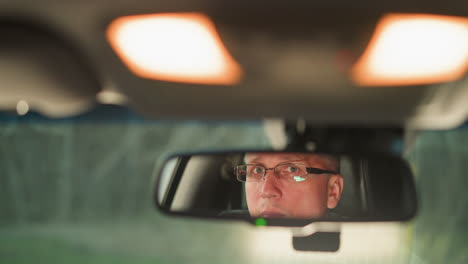 a man wearing glasses is reflected in the rearview mirror of a car. his expression is focused and contemplative, as the soft interior lighting illuminates the scene