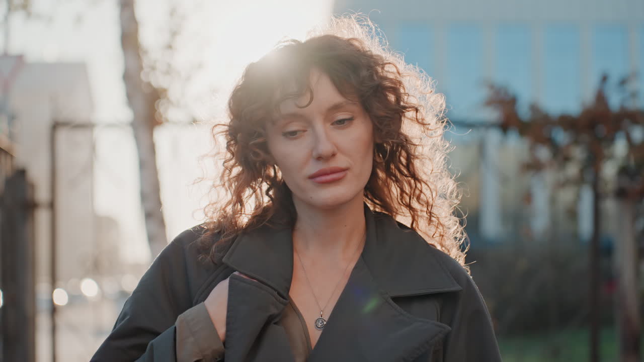 Recinzione all'ora d'oro, fiorista che cammina. Passeggiata cinematografica sul marciapiede con morbida retroilluminazione, capelli ricci contornati dal sole, espressione pensierosa mentre la fiorista lascia il negozio per una pausa, cancello in ferro ed edifici cittadini.