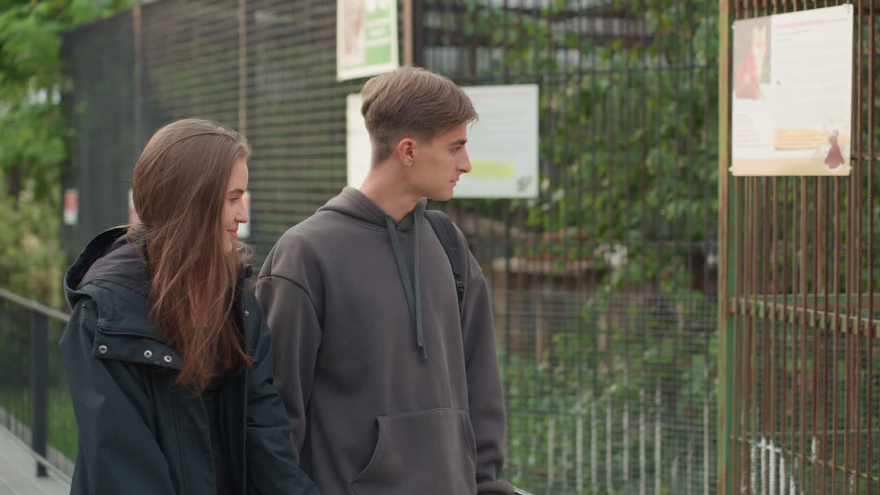 teenage couple walking in green park, casual jackets and hoodies, quiet conversation, leafy pathway beside metal fence, informational signs, relaxed pacing and candid smiles, study break atmosphere