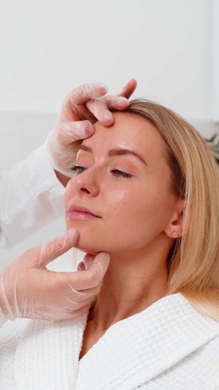 Cosmetologa con mascherina che esamina la pelle del viso di una giovane cliente prima di un trattamento estetico in clinica.