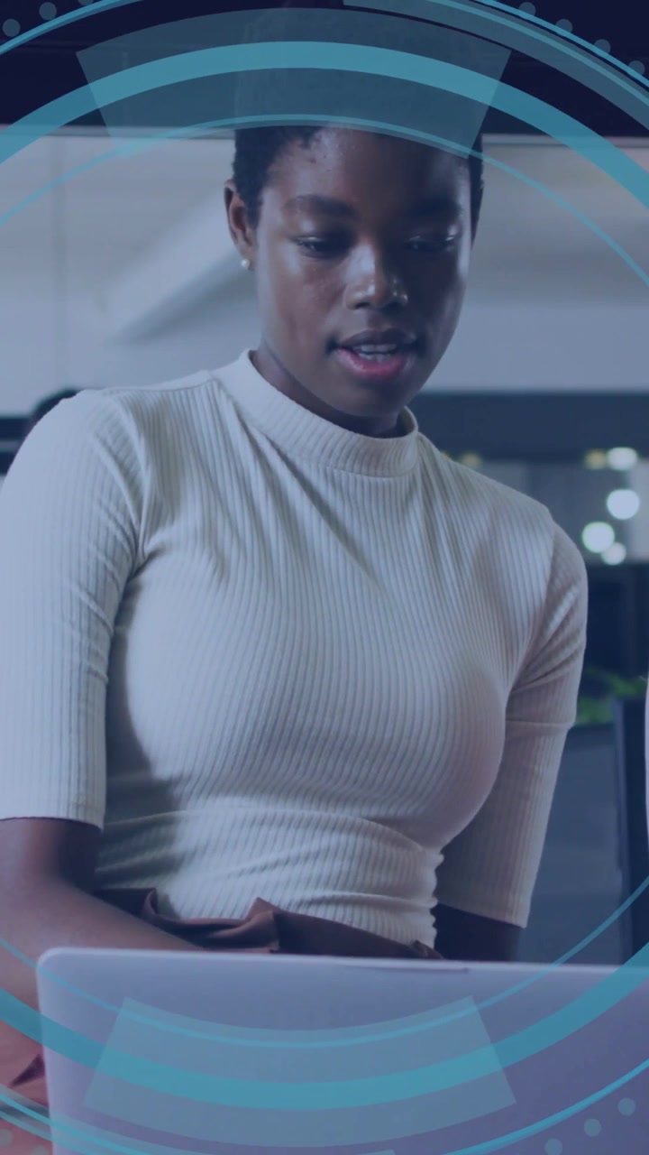 Vertical video: Woman leaning in and typing in office, tech HUD fading over laptop, analyzing data