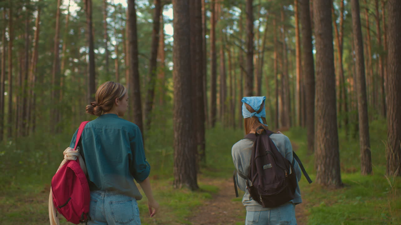 los primos caminan por un sendero forestal rodeado de exuberante vegetación, una prima balancea juguetonamente su bolsa negra sobre su hombro, mientras que la otra, vestida con una camisa verde, observa con una sonrisa